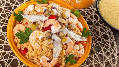 Un tavolo allestito con un piatto di couscous ai frutti di mare, immerso in un contesto estivo con sole e mare sullo sfondo.